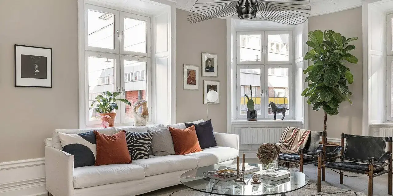 Vardagsrummet är ljust med en vit soffa, kuddar i olika färger och mönster, ett runt soffbord och en stor taklampa. En stor grön växt bidrar till trivseln.