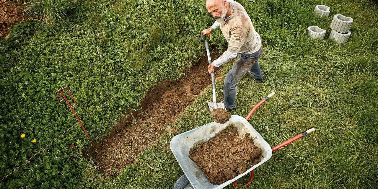 Mann gräbt einen Graben, um den Hang abzufangen. Erde wird in eine Schubkarre geschaufelt.