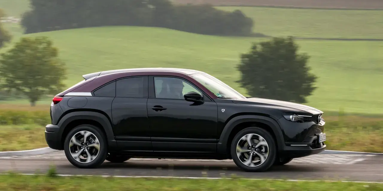 Czarna Mazda MX-30 z bordowym dachem sunie po drodze wśród zielonych wzgórz.