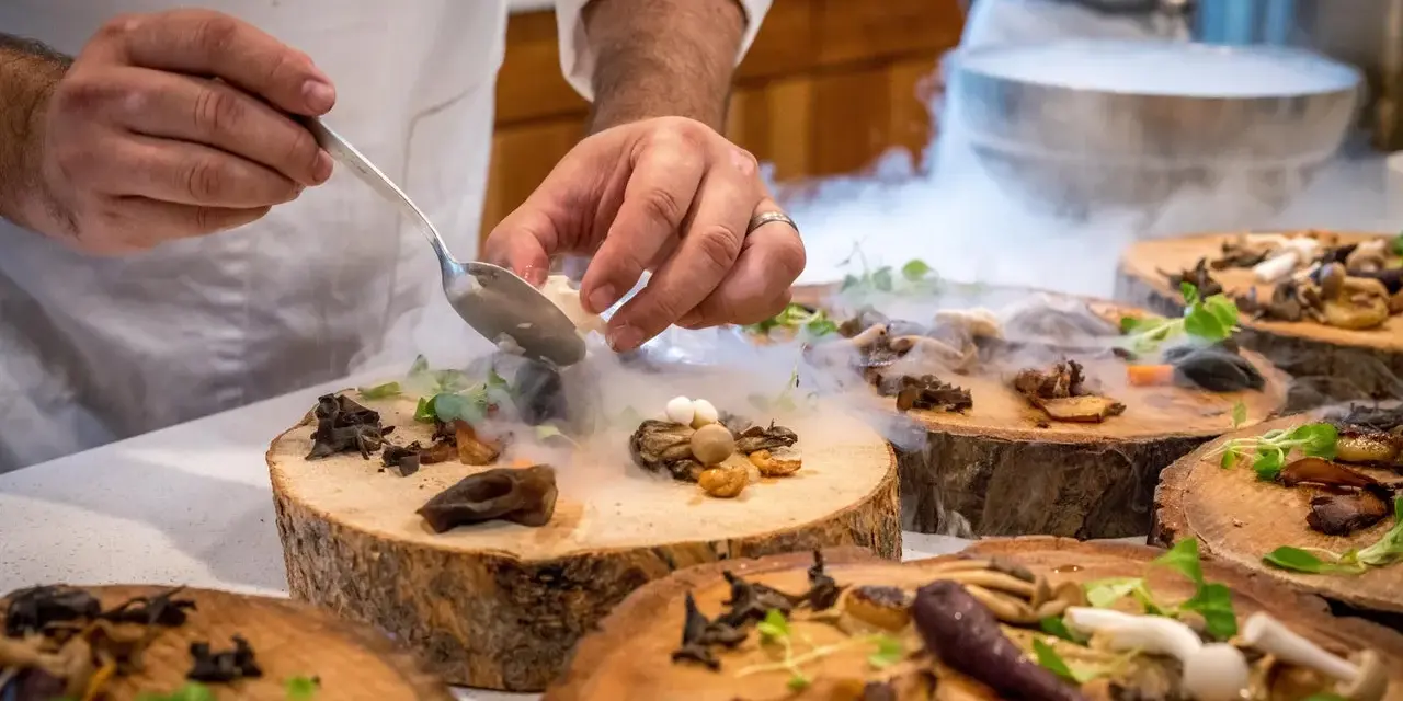 Szef kuchni serwuje danie z grzybami i ziołami na drewnianych plastrach, otoczone mgłą. To przykład nietypowych restauracji w Warszawie.