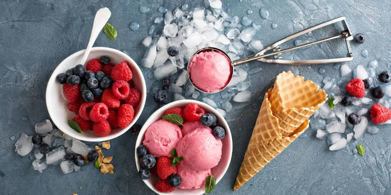 Himbeeren und Blaubeeren neben Schalen mit leckerem Milcheis selber machen. Eiswaffeln und ein Eisportionierer liegen bereit.