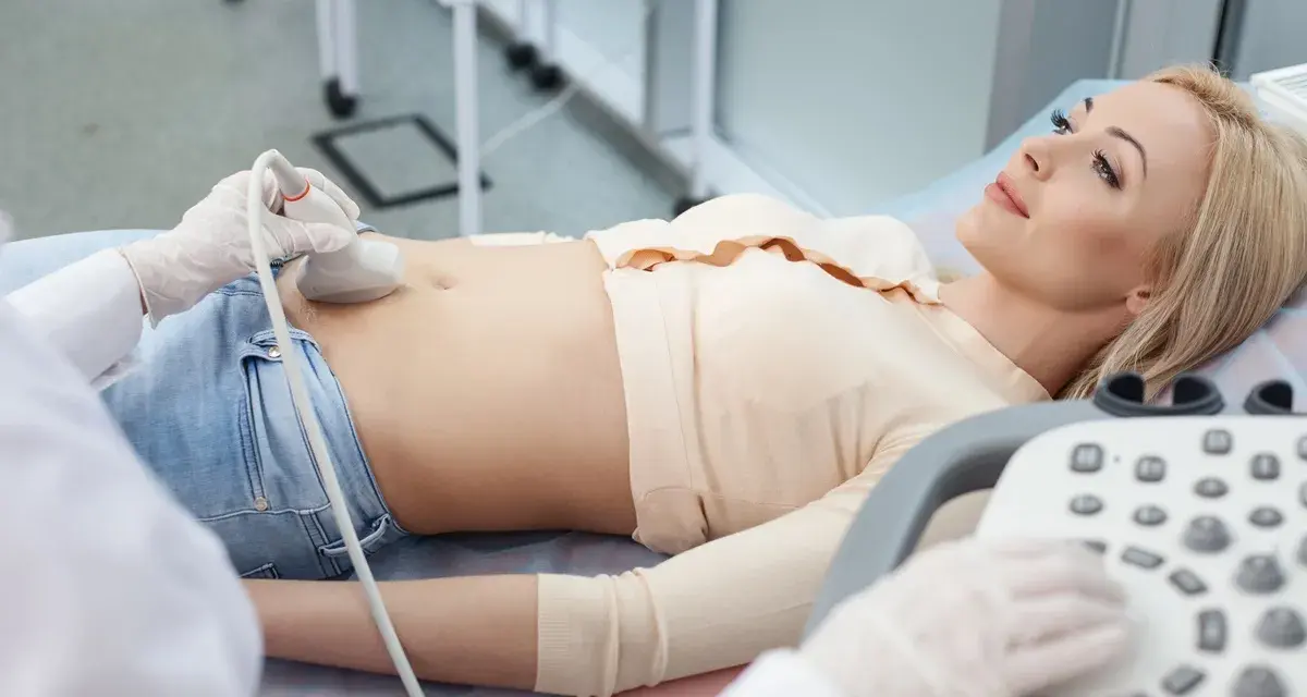 Kobieta leży na kozetce, a lekarz wykonuje jej usg 1 trymestru. Widoczna jest sonda aparatu USG przykładana do brzucha pacjentki.