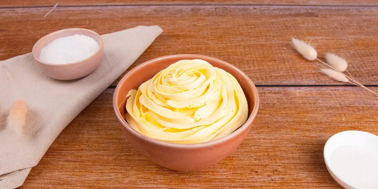 Una ciotola piena di soffice crema al burro, con zucchero in due piccole ciotole e decorazioni di erba secca su un tavolo di legno.