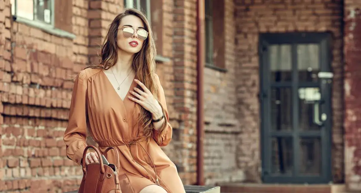Elegancka kobieta w brązowej sukience i okularach przeciwsłonecznych, z torebką. Idealne buty do brązowej sukienki podkreślają jej styl.