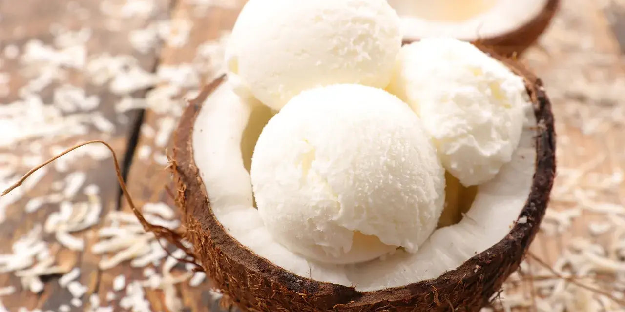 Cremiges Kokosnuss Eis in einer halbierten Kokosnuss, umgeben von Kokosraspeln auf Holz. Ein tropischer Genuss!