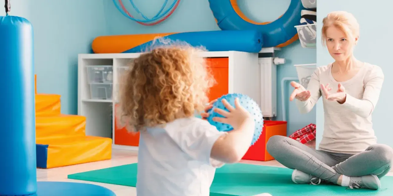 Ergotherapie für Kinder: Eine Therapeutin leitet ein Kind mit lockigem Haar an, das einen blauen Ball hält, in einem bunten Raum voller Spielgeräte.