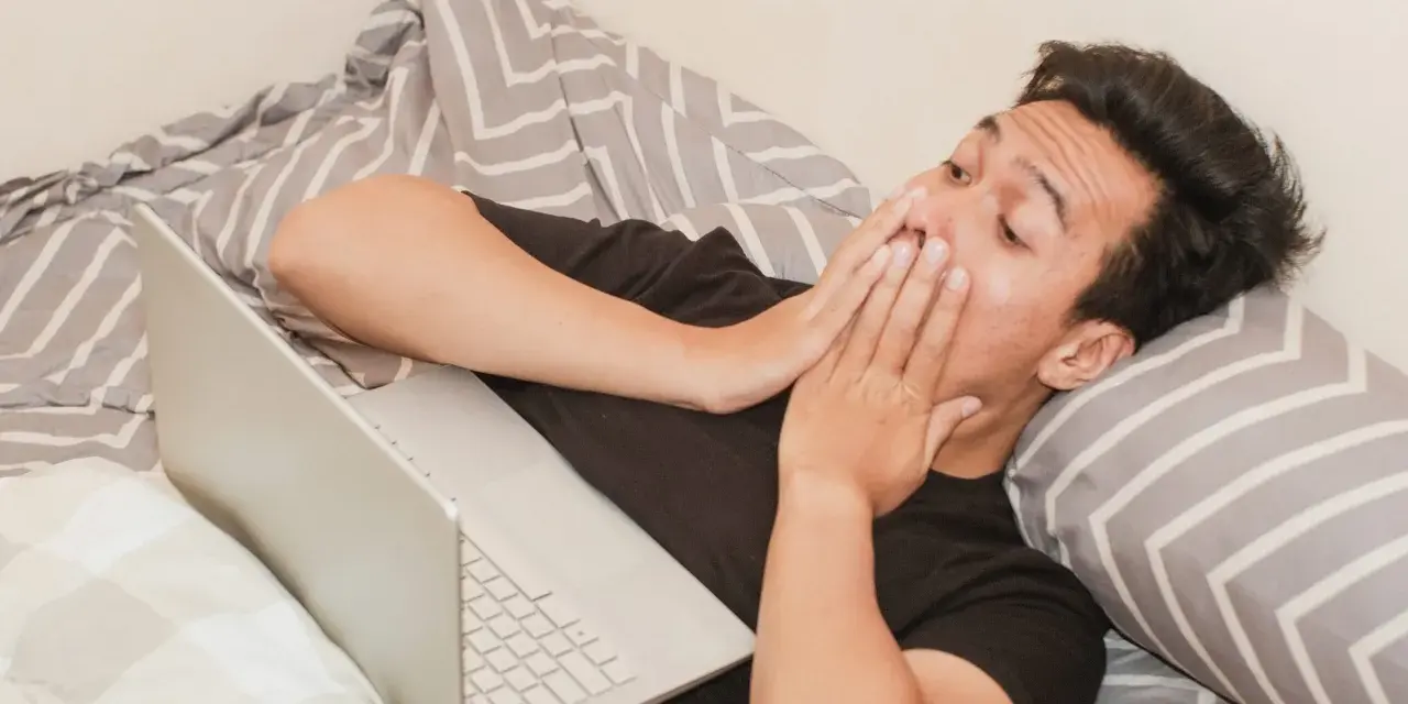 Mężczyzna w łóżku z laptopem, zakrywający usta dłońmi, może symbolizować walkę z uzależnieniem od pornografii.