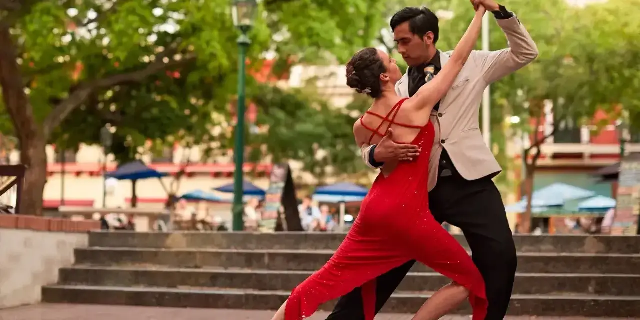 Para tańczy tango, latynoamerykański taniec towarzyski, w czerwonej sukni i eleganckim garniturze, na tle miejskiego placu.