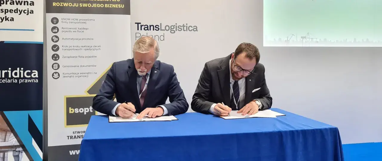 Dwóch mężczyzn w garniturach podpisuje dokumenty. Obok baner z napisem TransLogistica Poland, sugerujący umowę w branży gpw logistics.