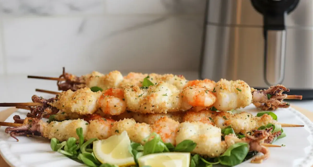 Spiedini di gamberi e calamari impanati, cotti alla perfezione nella friggitrice ad aria, serviti su un letto di insalata con spicchi di limone.
