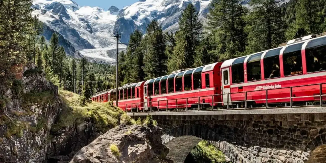 A red train travels across a stone bridge, winding its way from Chur to Tirano through a stunning alpine landscape with snow-capped mountains.