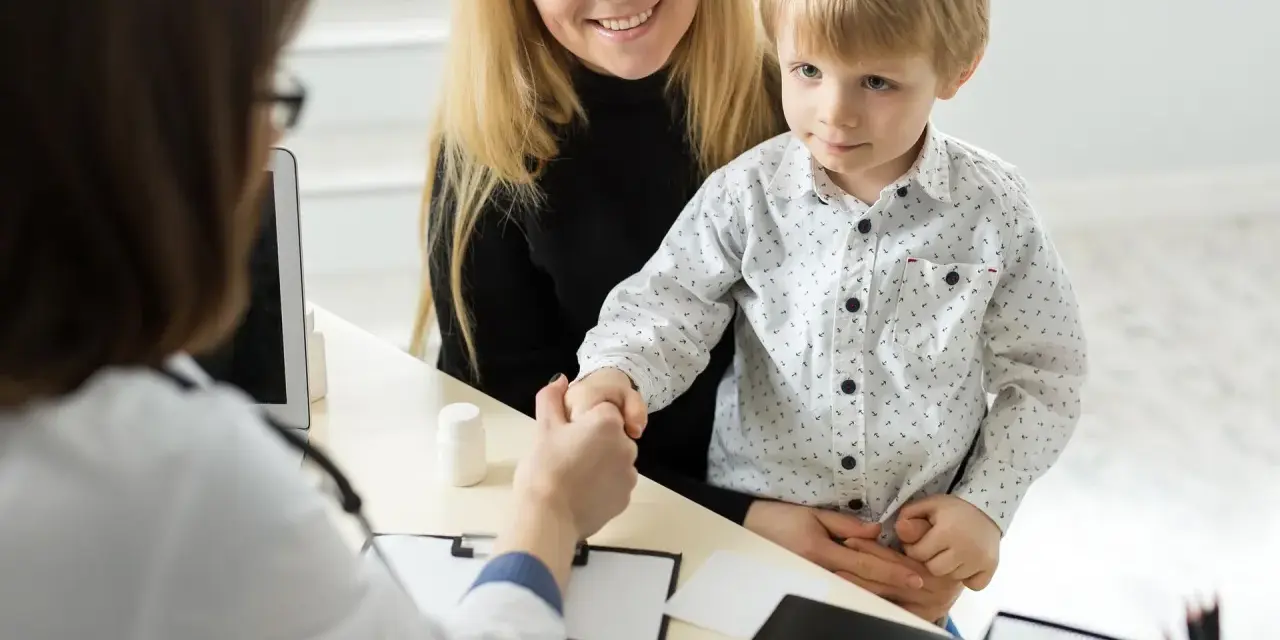 Mama z synkiem u lekarza. Chłopiec ściska dłoń lekarce, a mama uśmiecha się. W tle widać tablet i białą buteleczkę, jakby z lekiem. To ważny moment, być może wpis do książeczki zdrowia.