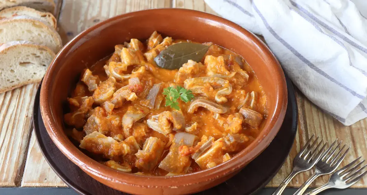 Guiso casero de oreja de cerdo en salsa de tomate, servido en cazuela de barro, acompañado de pan rústico y tenedores.