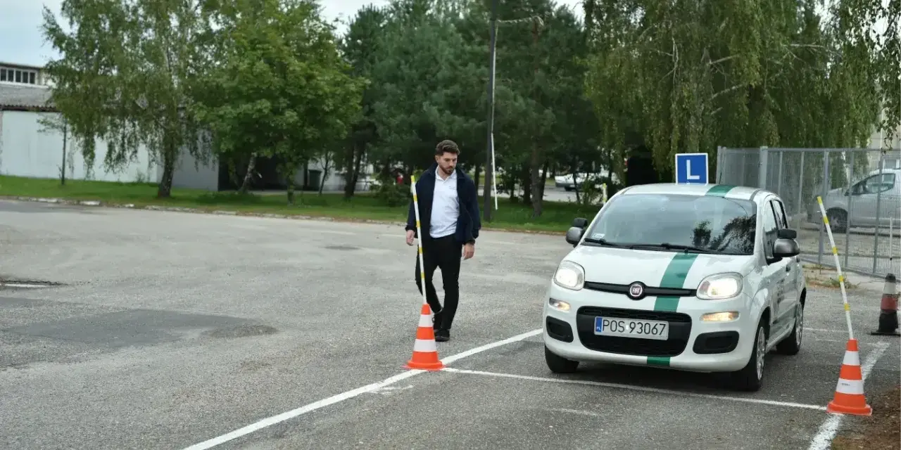 Młody mężczyzna ustawia pachołek na placu manewrowym. Biały Fiat Panda z literą "L" na dachu czeka na swój egzamin praktyczny kat b.