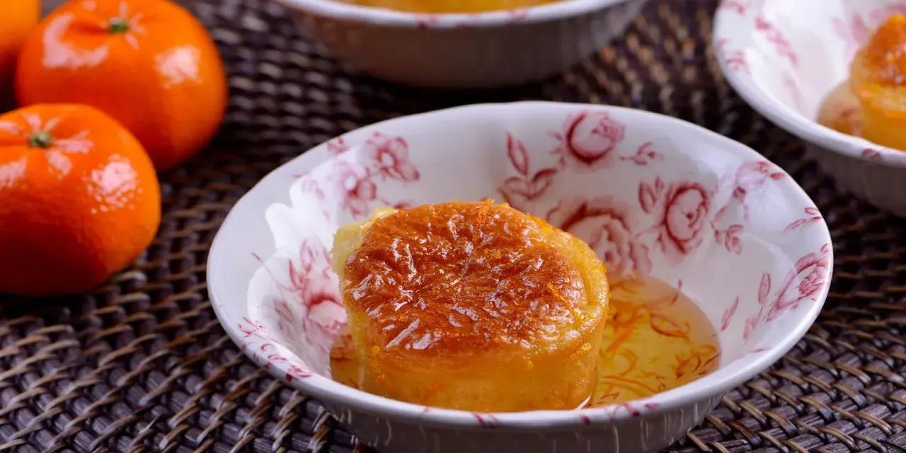 Un esponjoso baba al ron bañado en almíbar, adornado con ralladura de naranja, en un cuenco floral. Cerca, mandarinas frescas.