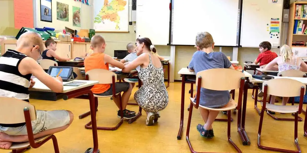 Dzieci siedzą w klasie, ucząc się. Nauczycielka pochyla się nad jednym z uczniów. Ile jest klas w Anglii? Tego nie wiemy, ale ta lekcja wygląda na interesującą.