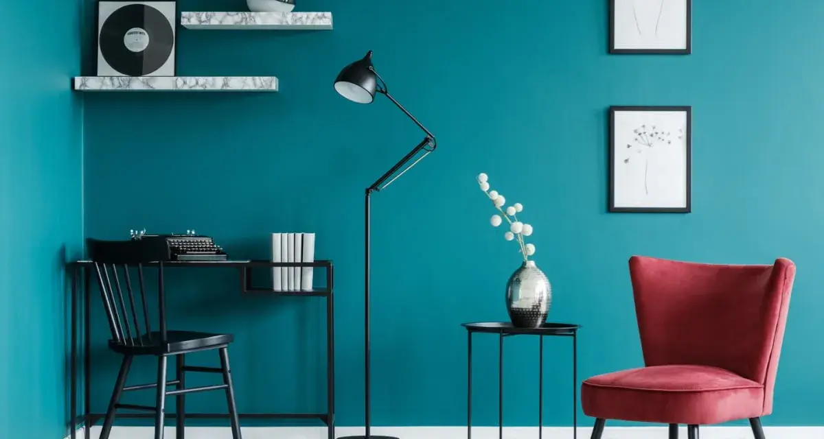 Angolo studio con pareti color **blu pavone**, scrivania nera, sedia, lampada e poltroncina rossa.