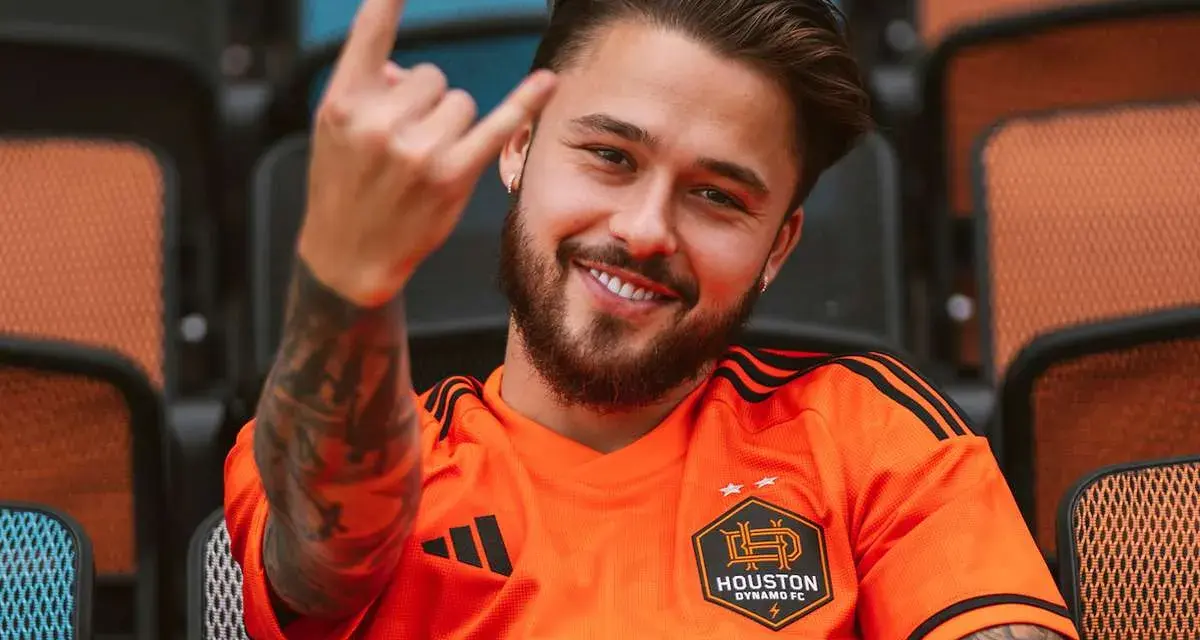 Uśmiechnięty Adam Kowalczyk w koszulce Houston Dynamo FC pozdrawia gestem rock and rolla, siedząc na trybunach.
