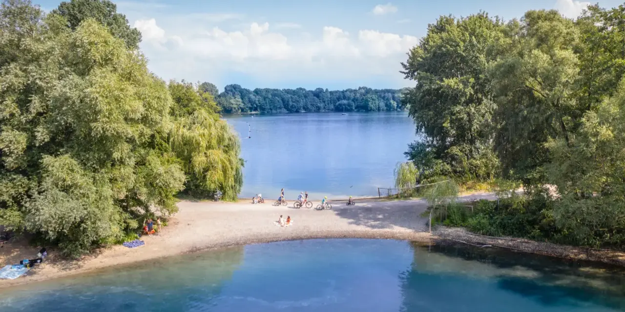 Sommerliche Idylle am Strandbad Wedau: Menschen genießen den Tag am Seeufer, einige radeln, andere entspannen im Sand.