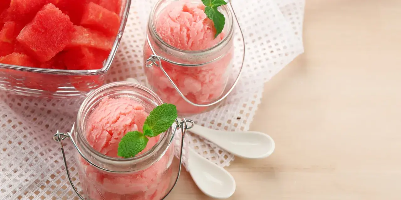 Erfrischendes Wassermelonen-Eis in Gläsern, garniert mit Minze. Daneben ein Schälchen mit frischen Wassermelonenstücken.