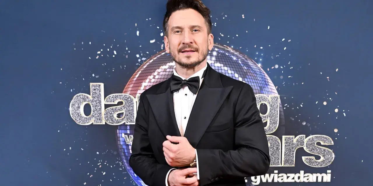 Tomasz Barański w czarnym smokingu pozuje na tle błyszczącej kuli dyskotekowej i napisu "Dancing with the Stars".