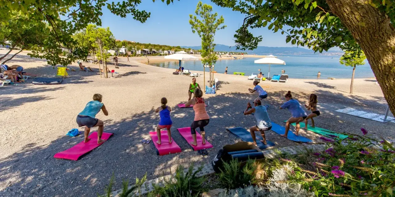 Grupa osób ćwiczy jogę na plaży w Omisalj. W tle widać morze i kemping.