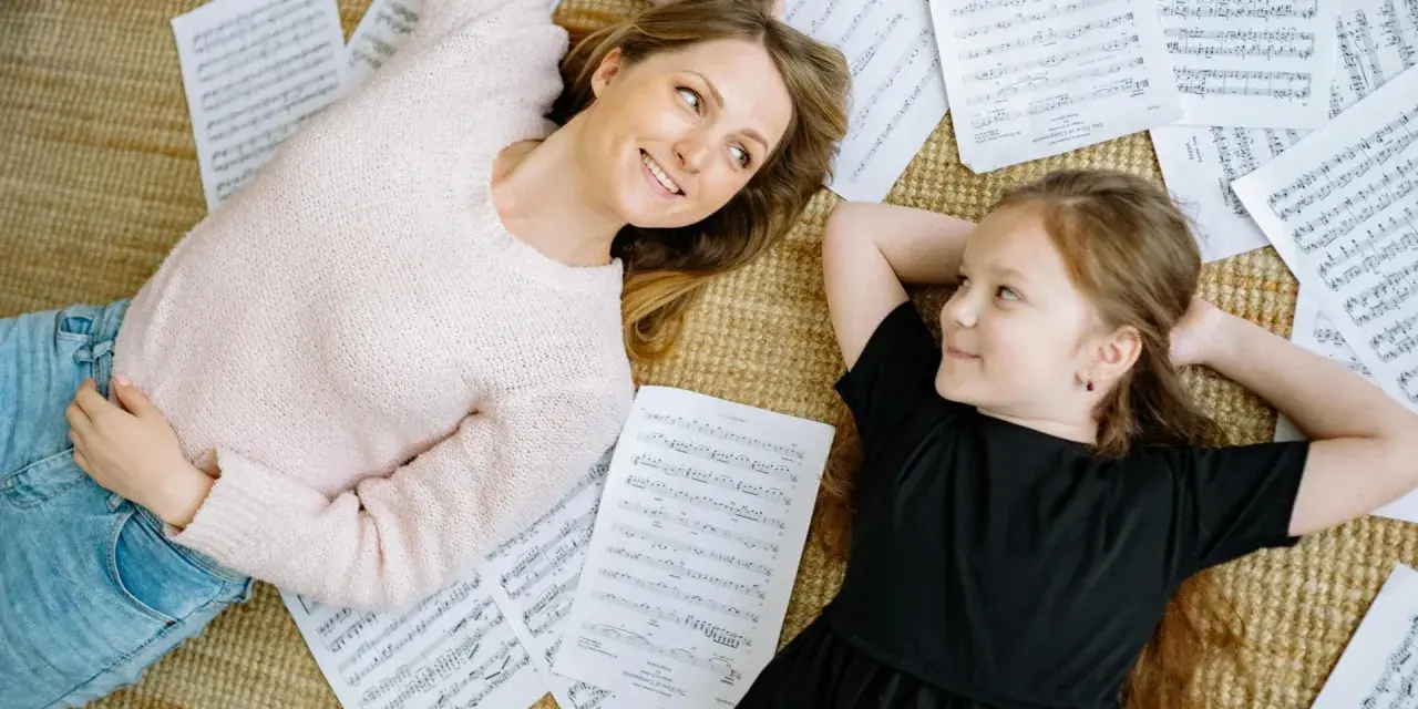 Mutter und Tochter liegen auf Notenblättern und lächeln sich an. Ein friedlicher Moment, der zeigt, wie Musik im Alltag verbindet.
