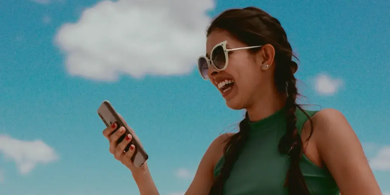 Lachende Frau mit Sonnenbrille hält Smartphone vor blauem Himmel mit Wolken.
