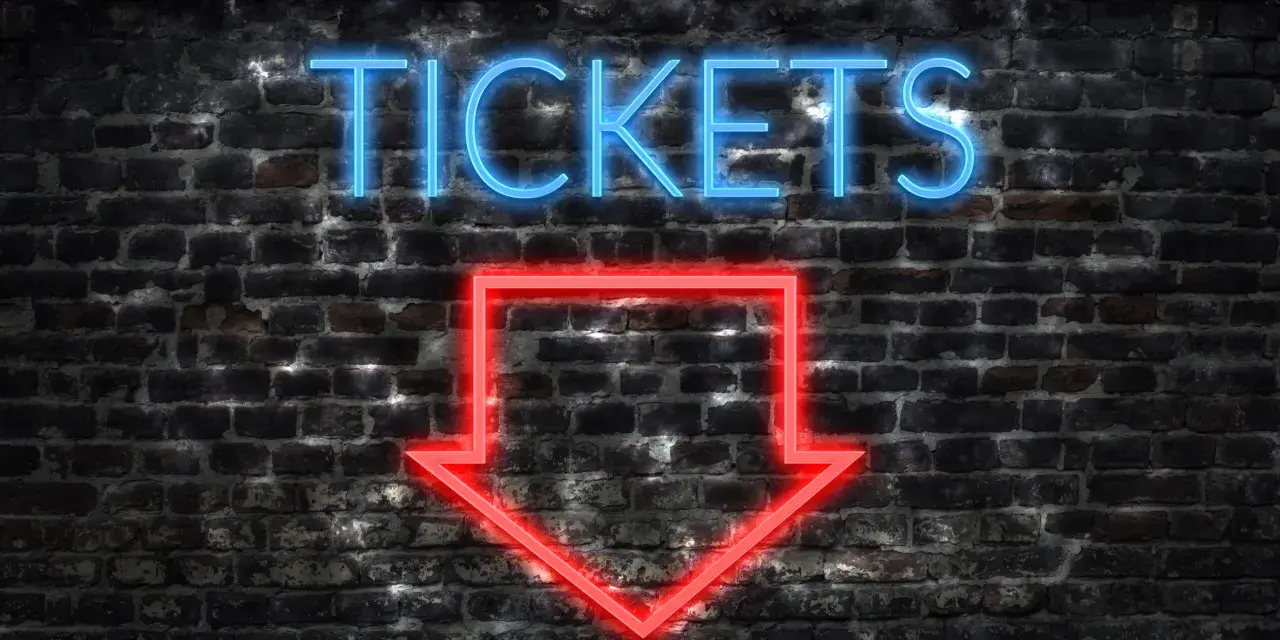 Napis "TICKETS" neonem w kolorze niebieskim i czerwona strzałka w dół na ceglanej ścianie.