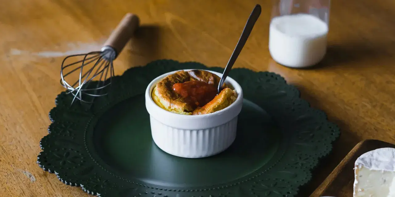 Delicioso souffle de vainilla con salsa de fresa, listo para disfrutar. A su lado, un batidor y un vaso de leche.