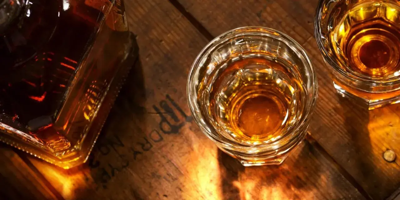 Butelka i dwa kieliszki z bursztynowym płynem, przypominającym koniak lub whisky, stoją na drewnianym blacie.