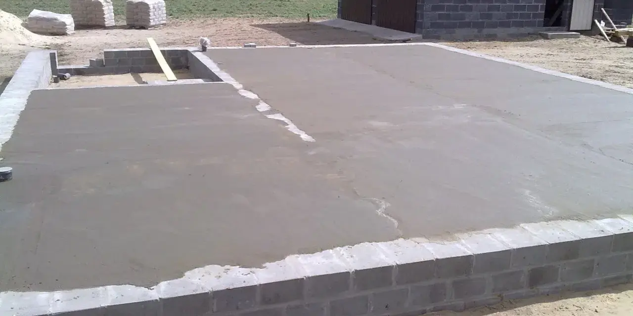 Świeżo wylany beton na chudziaka tworzy fundament pod budowę. W tle widać materiały budowlane i fragment budynku.