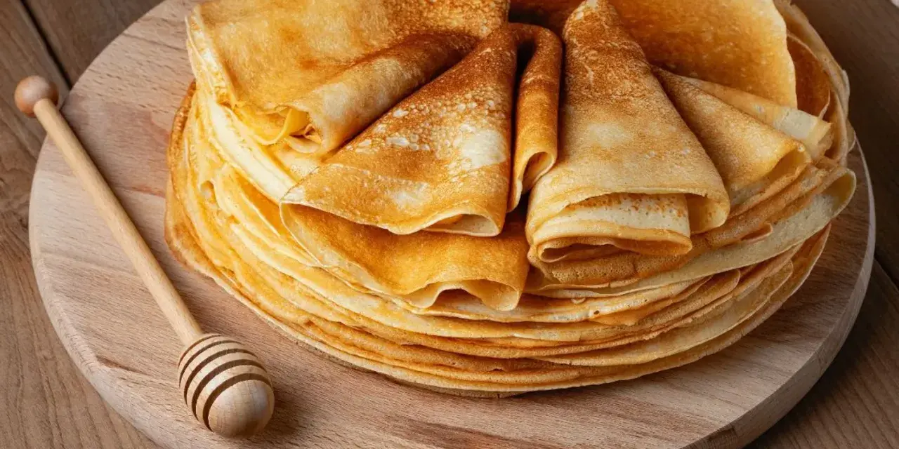Pile de crêpes dorées, prêtes à être garnies. Une cuillère à miel en bois repose à côté, suggérant une délicieuse recette de crêpes.