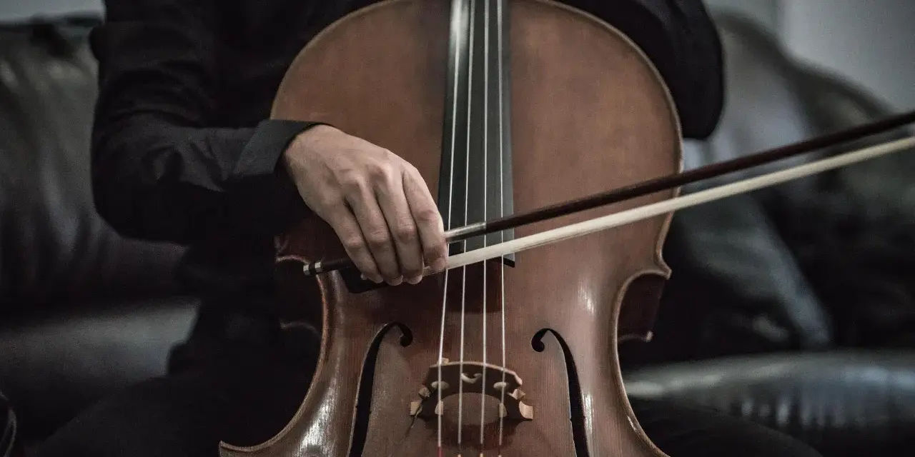 Eine Hand hält den Bogen über dem Violoncello-Instrument, bereit, die Saiten zum Klingen zu bringen.