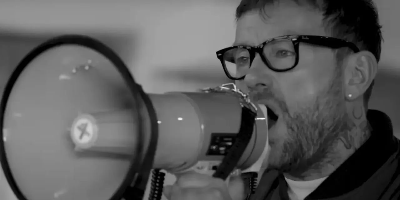 Un homme avec des lunettes et un mégaphone, sa voix résonne. Le groupe est en plein concert.