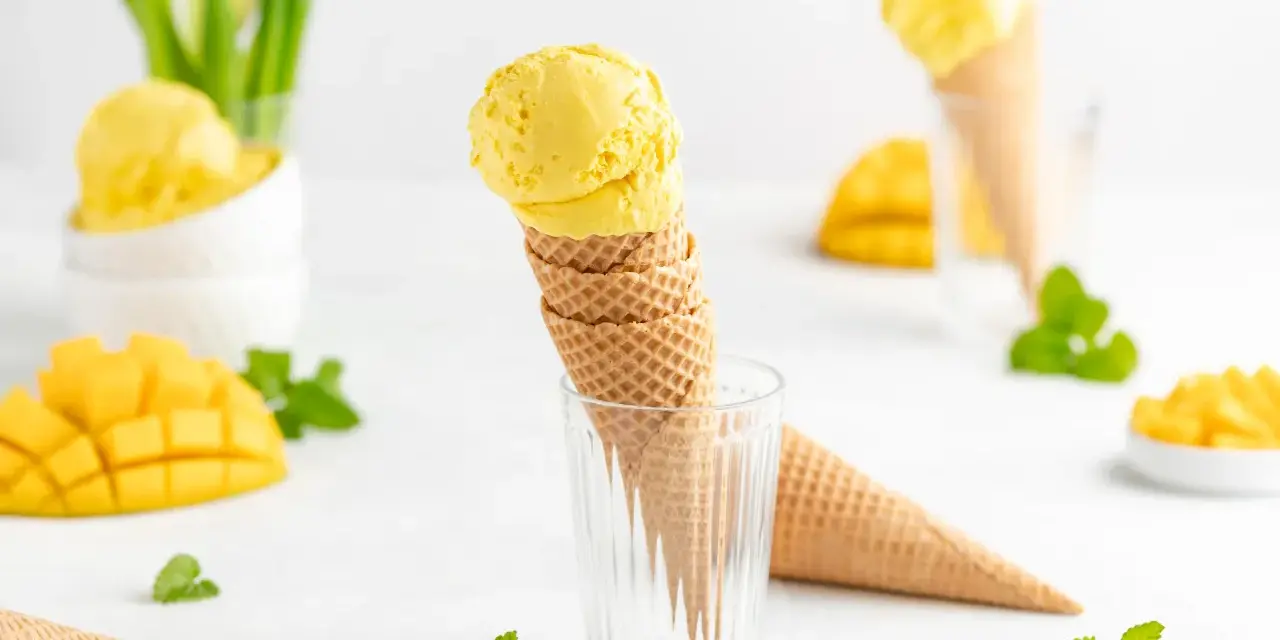 Zwei Kugeln cremiges Mango Eis in Waffelhörnchen, umgeben von frischen Mangostücken und Minze. Ein süßer Genuss!