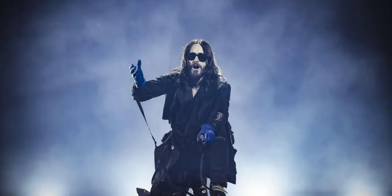 Jared Leto w niebieskich rękawiczkach, w okularach i długim płaszczu, klęczy na scenie. Jego zespół pewnie czeka na jego znak.