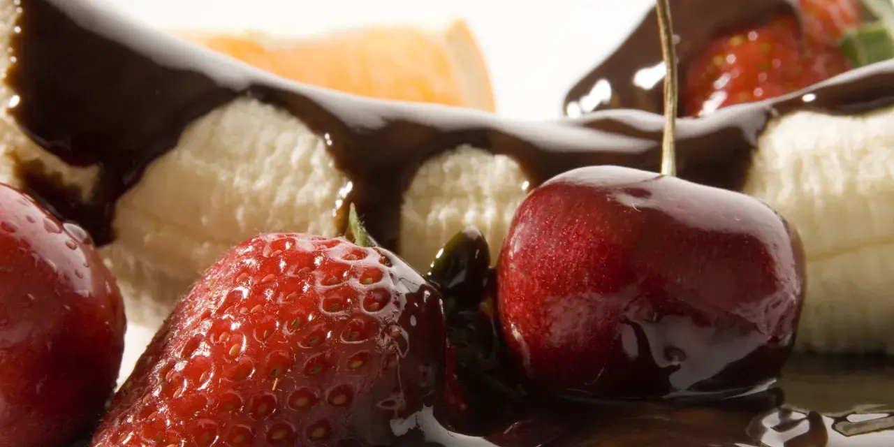 Soczyste truskawki i wiśnie oblane czekoladą, obok kawałków banana. Pyszne owoce w czekoladzie kuszą swoim wyglądem.