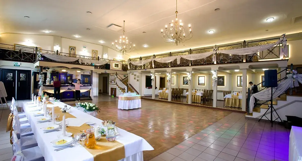 Elegancka sala weselna z długim stołem, ozdobionym złotą podkową. W tle bar i schody prowadzące na antresolę.