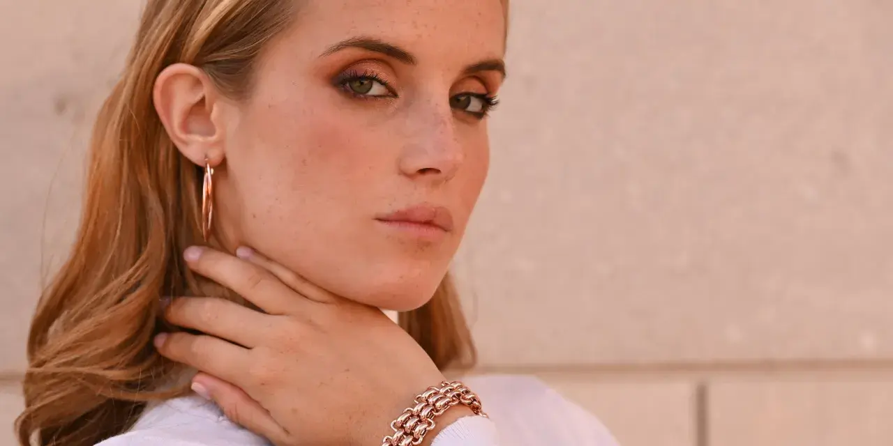Donna con lentiggini e gioielli in oro rosa, un bracciale a maglie larghe e orecchini a cerchio.