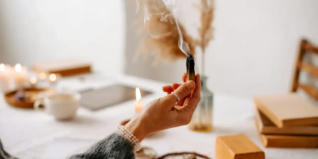 Mano sosteniendo un palo santo humeante, rodeada de velas, libros y cristales, creando un ambiente de sanación y paz.