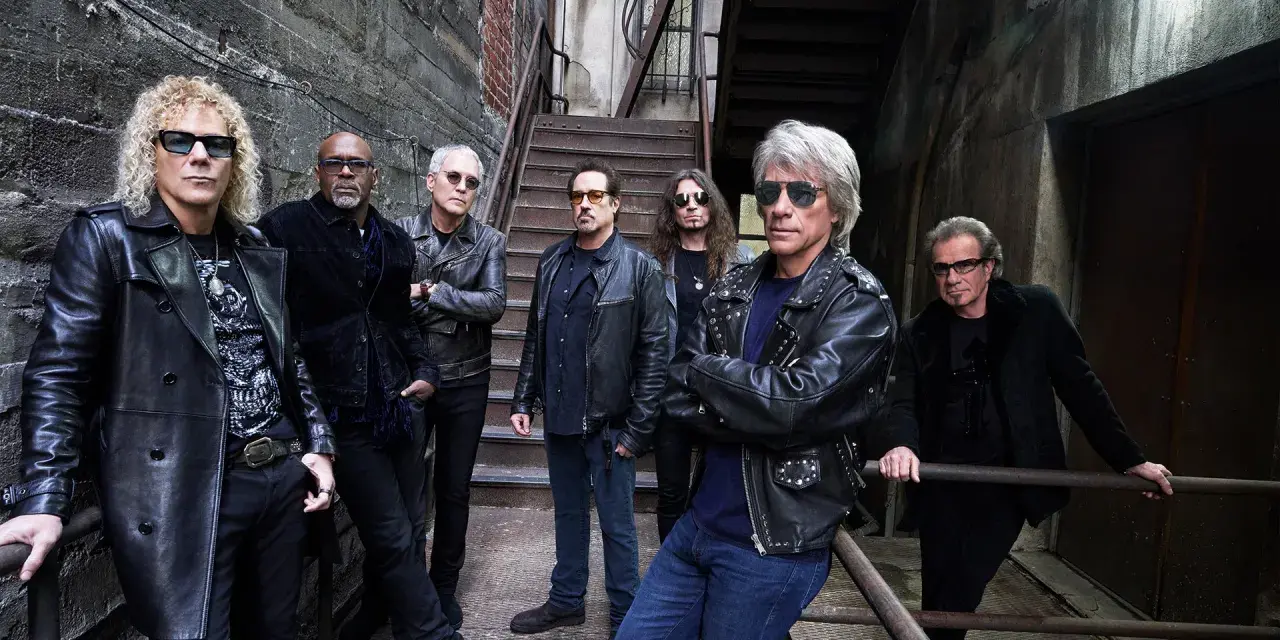 Zespół Bon Jovi gotowy na koncert, w skórzanych kurtkach i okularach przeciwsłonecznych, pozuje na tle industrialnych schodów.