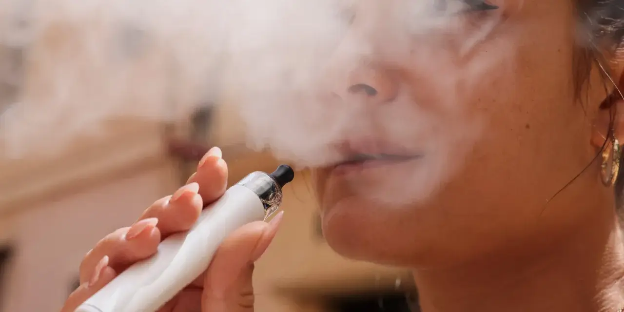 Una mujer exhala vapor de un vaper blanco. El vapor parece rodear su rostro, sugiriendo que el vaper encharca los pulmones.