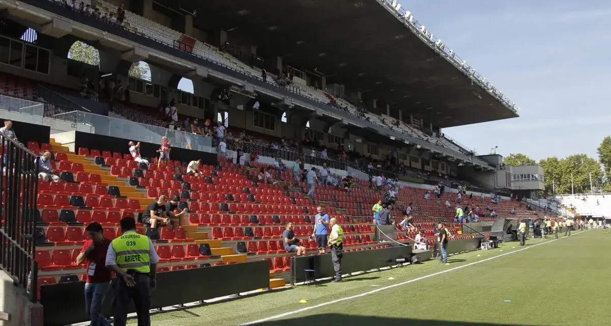Puste trybuny stadionu Vallecas, z kilkoma osobami siedzącymi i stojącymi.