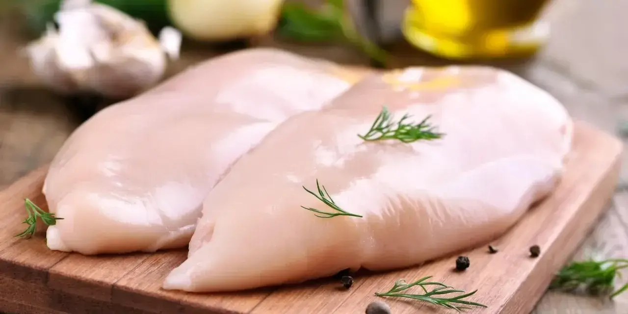 Dwie surowe piersi z kurczaka na desce, obok czosnek, cebula i koperek. Zastanawiasz się, ile białka ma pierś z kurczaka?