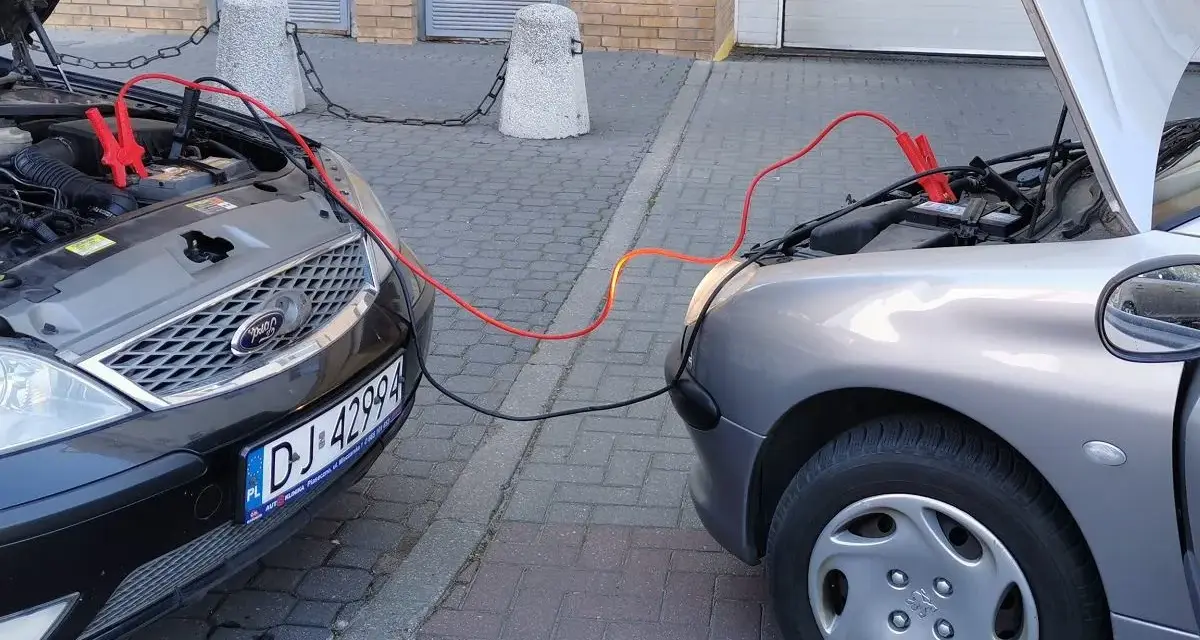 Dwa samochody z otwartymi maskami, połączone kablami rozruchowymi. Pomoc w awaryjnej sytuacji, jak sprawdzić masę w aucie.