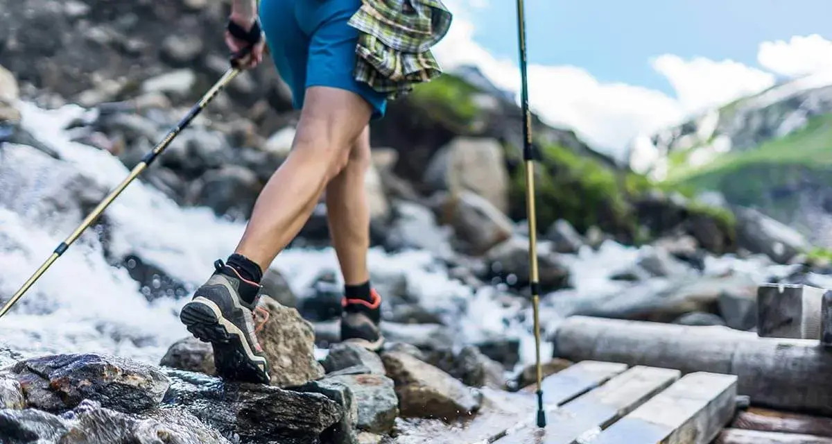 Une personne en tenue de ppg sport traverse un ruisseau sur un pont de bois, bâtons de randonnée en main. Paysage de montagne.