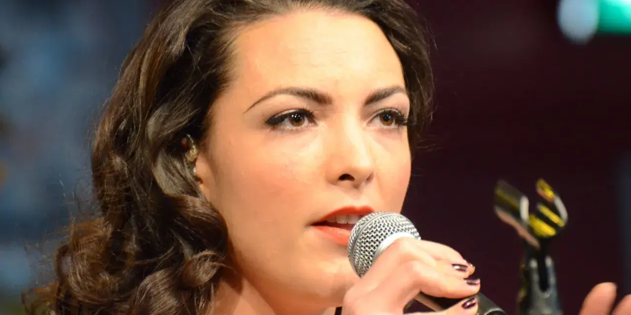 Caro Emerald śpiewa do mikrofonu, zbliżenie na twarz i dłonie.