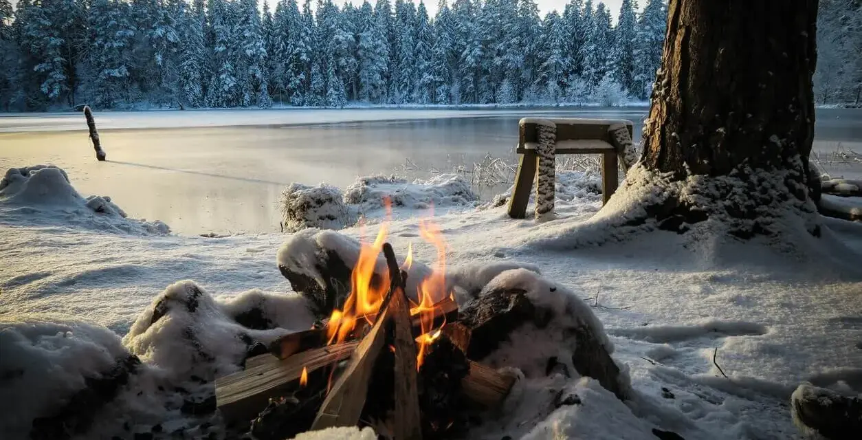 Ein Lagerfeuer wärmt beim Zelten im Winter am gefrorenen See. Schnee bedeckt die Ufer und den Wald im Hintergrund.