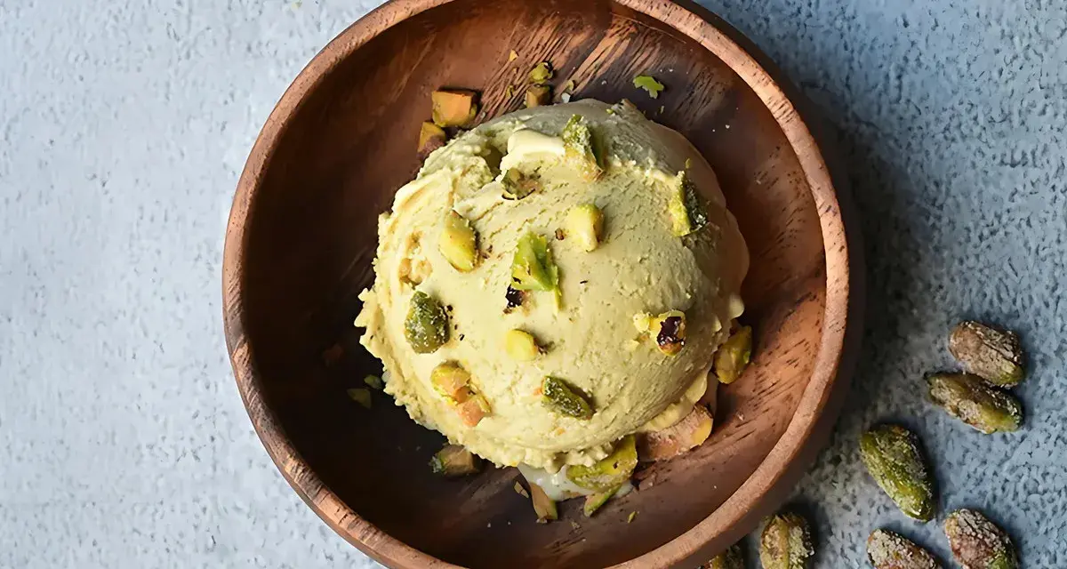 Cremoso helado de pistacho servido en un cuenco de madera, adornado con trozos de pistacho y acompañado de más pistachos enteros esparcidos alrededor.
